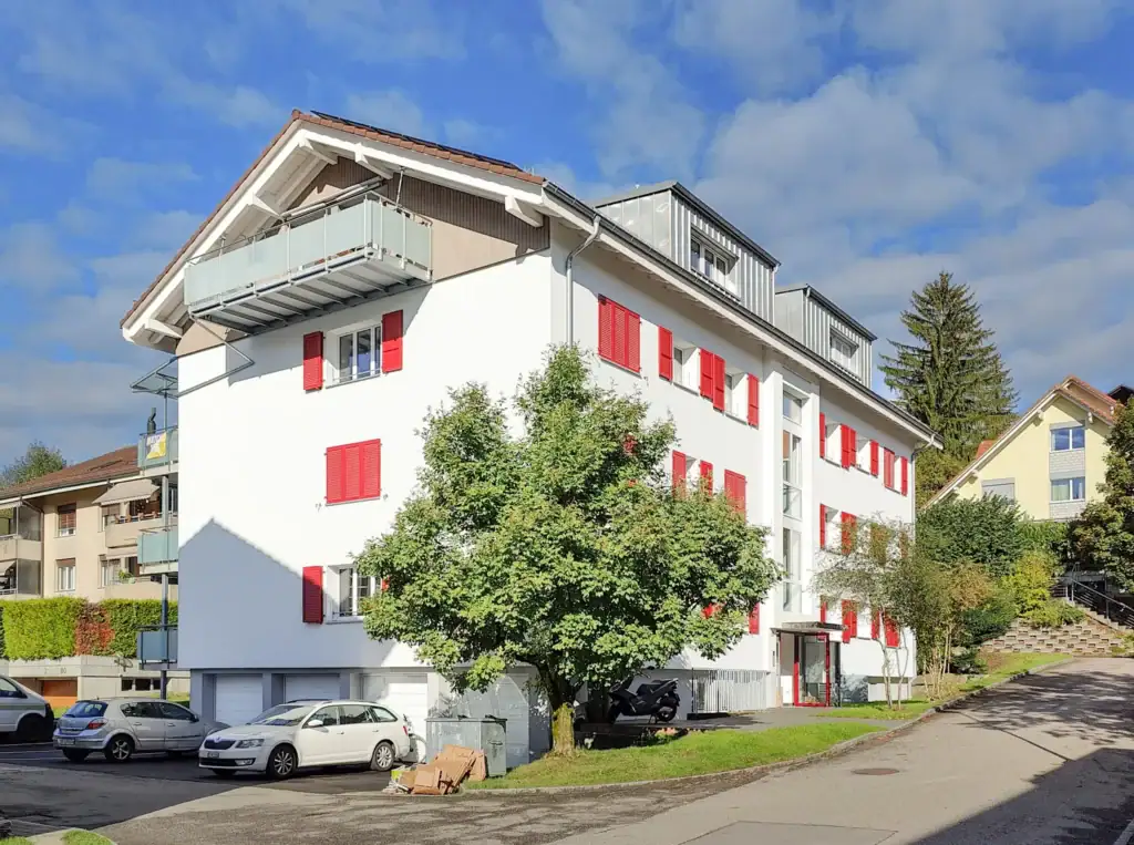 Mehrfamilienhaus nach Sanierung mit klar gegliederter Fassade, roten Fensterläden und ausgebautem Dachgeschoss, ruhige Wohnlage mit Aussenraum und Parkierung.