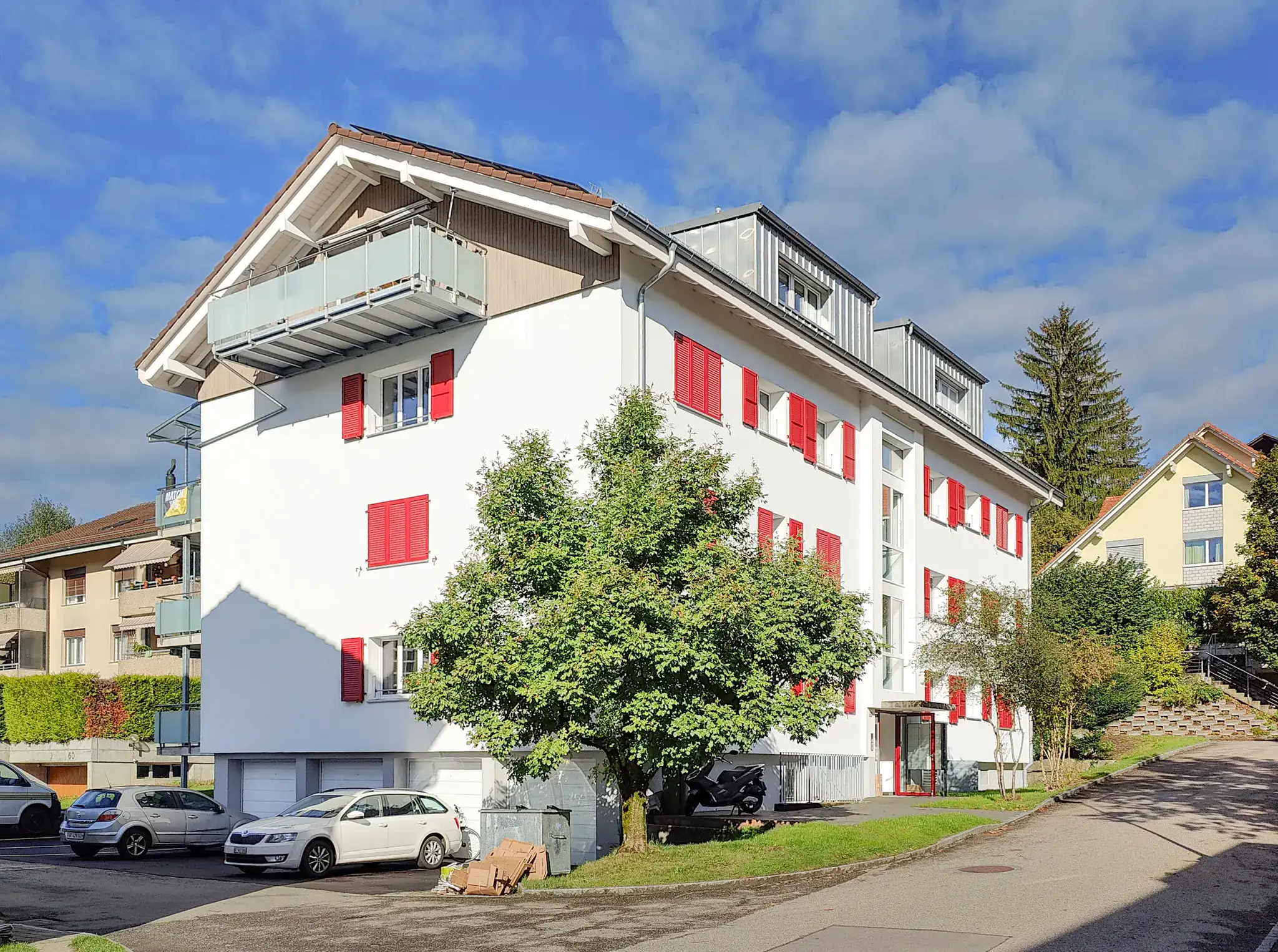 Mehrfamilienhaus nach Sanierung mit klar gegliederter Fassade, roten Fensterläden und ausgebautem Dachgeschoss, ruhige Wohnlage mit Aussenraum und Parkierung.