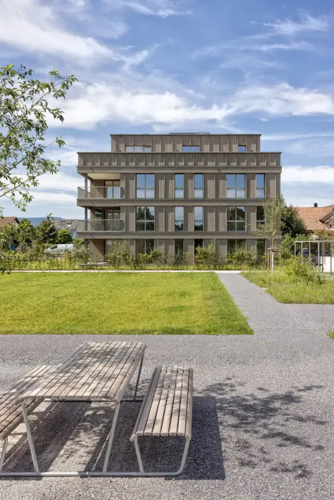 Modernes Mehrfamilienhaus mit Holzfassade, grosszügigem Grünraum und gemeinschaftlichem Aussenbereich.